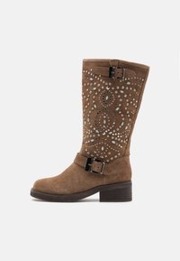 Bota hasta la rodilla de ante marrón con tachuelas decorativas y un patrón de remolinos único. Incluye una correa ancha y un detalle de hebilla metálica.