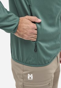 Grüne Reißverschlussjacke aus strukturiertem Stoff, getragen über hellbeigen Hosen mit einer Seitentasche und Logo. Der Reißverschlussanhänger hat eine schwarze Schnur.