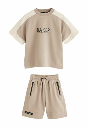 Boy walking indoors wearing a beige short-sleeve shirt with "BAKER" text and matching shorts, white sneakers, and striped socks.