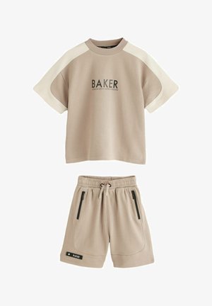 Boy walking indoors wearing a beige short-sleeve shirt with "BAKER" text and matching shorts, white sneakers, and striped socks.