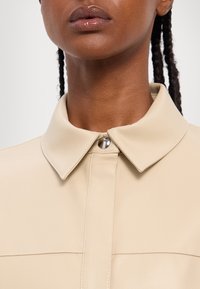 Beige leren shirt met een klassieke kraag, drukknopen sluiting en zichtbare stikdetails aan de voorkant. Glad textuur.