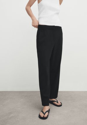 Personne portant un pantalon noir sur mesure, un haut blanc sans manches et des tongs noires, ajustant la taille sur un sol gris uni et un mur blanc.