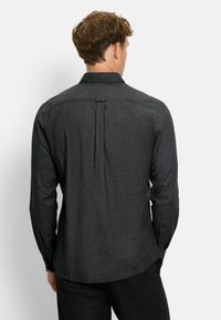 Mann mit kurzem braunem Haar, der ein dunkelgraues Langarmshirt und schwarze Hosen trägt, von hinten vor einem schlichten Hintergrund gesehen.