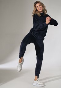 Marineblauw velours trainingspak met een langemouwen top en aansluitende joggingbroek. Gecombineerd met witte sportsneakers. Glad textuur in het hele pak.