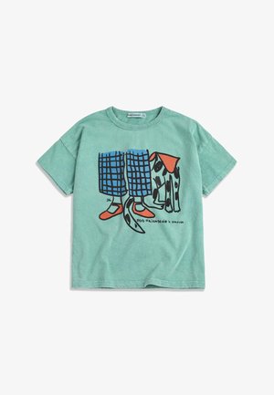 Mätovo zelené tričko s ilustráciou nôh v modrých károvaných nohaviciach a červených topánkach stojacich vedľa škvrnitého psa s červeným šálom.