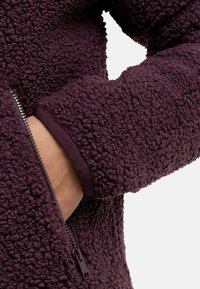 Veste en polaire de couleur violet foncé avec une surface texturée. Comprend une poche zippée et des poignets côtelés. Gros plan montrant la manche et l'accès à la main.