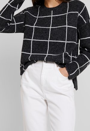 Pull gris foncé avec motif quadrillé blanc associé à un pantalon blanc taille haute, une main dans la poche, l'autre levée près de la tête.