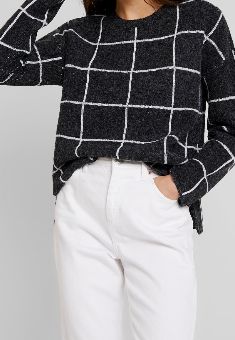 Pull gris foncé avec motif quadrillé blanc associé à un pantalon blanc taille haute, une main dans la poche, l'autre levée près de la tête.