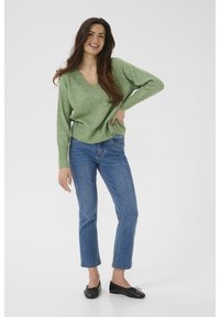 Pull vert à col en V en tissu tricoté doux, associé à un jean droit bleu clair et des ballerines noires, porté par une femme souriante.