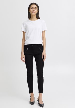 Donna in piedi con una maglietta bianca a maniche corte, pantaloni neri aderenti con tasche con zip e tacchi a punta neri, su uno sfondo bianco uniforme.