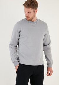Felpa grigia chiara con finitura testurizzata, scollo rotondo e maniche lunghe. Presenta un discreto dettaglio a forma di V sul colletto. Indossata con pantaloni scuri.