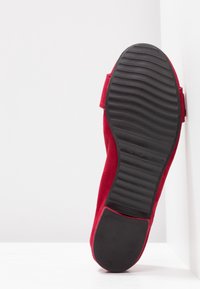 Semelle d'une chaussure plate rouge avec une semelle extérieure en caoutchouc noir texturé, montrée debout sur fond blanc.