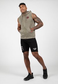 Mouwloze olijfgroene hoodie met een rits aan de voorkant en zijzakken, gecombineerd met zwarte shorts met bedrukte logo's en accenten. Zwarte sneakers.