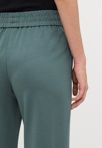 Ljusgröna byxor med elastisk midja och en slät textur. Närbild som visar en hand vilande på tyget, vilket framhäver detaljerna på midjebandet.