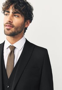 Sort jakkesæt med skræddersyet pasform, hvid skjorte og brunt tekstureret slips. Jakken har en diskret lomme. Blødt lys.