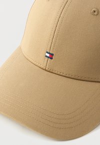 Cappellino da baseball beige con cuciture visibili e piccolo emblema rettangolare rosso, bianco e blu sul pannello anteriore.