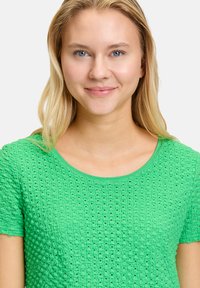 Grünes gestricktes T-Shirt mit kurzen Ärmeln, das ein strukturiertes Design mit kleinen Löchern und einem Rundhalsausschnitt aufweist. Hergestellt aus leichtem Stoff.