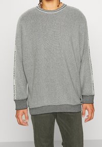 Persona che indossa un maglione grigio con texture e dettagli neri e loghi sulle maniche, abbinato a pantaloni verdi, in piedi davanti a uno sfondo bianco.