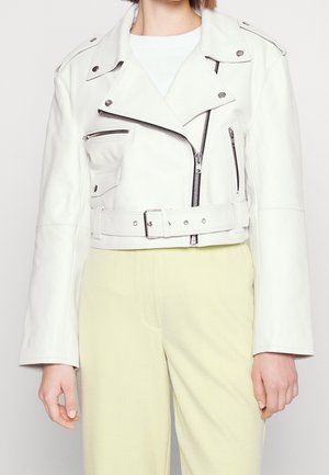 Femme portant une veste biker en cuir blanc avec des poches zippées et une taille ceinturée, assortie à un pantalon jaune clair.