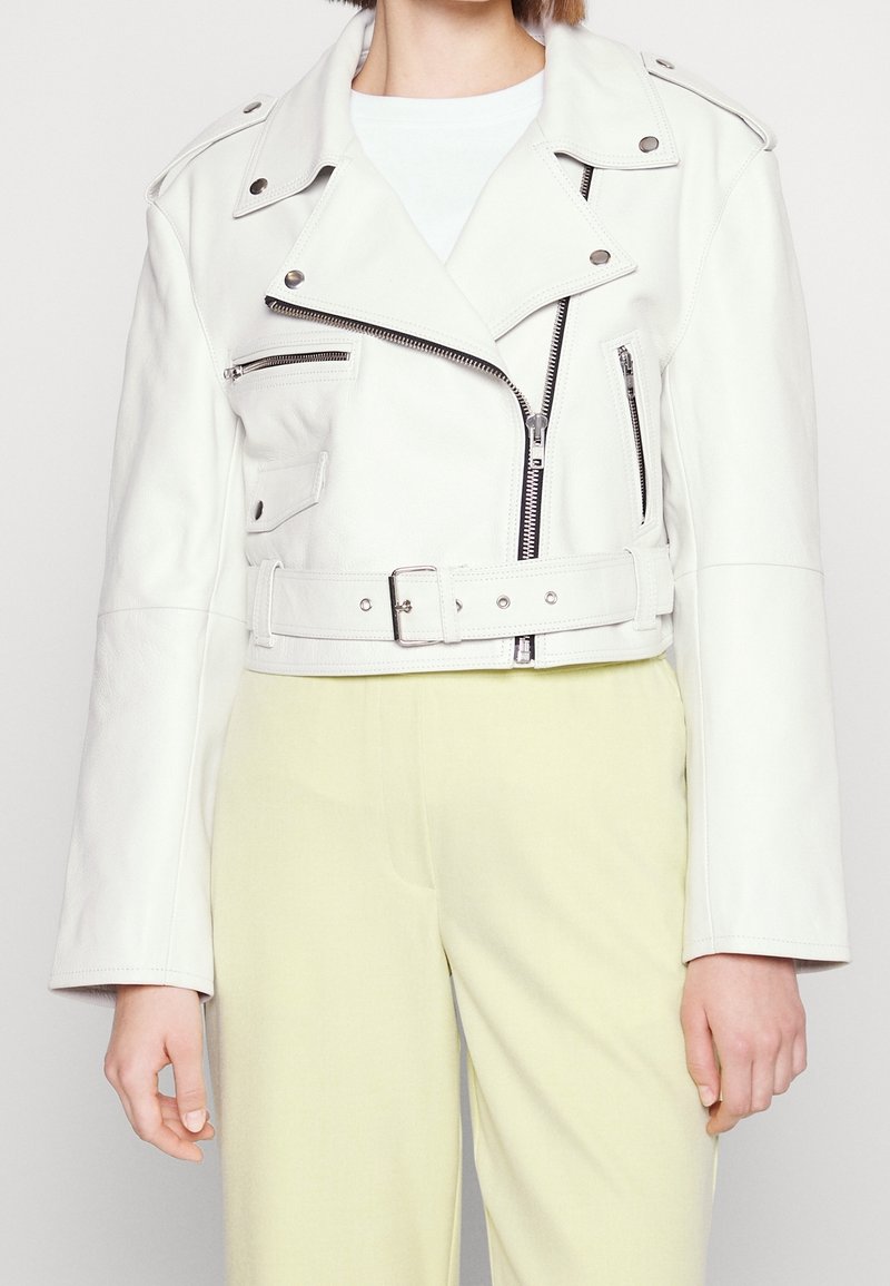 Femme portant une veste biker en cuir blanc avec des poches zippées et une taille ceinturée, assortie à un pantalon jaune clair.