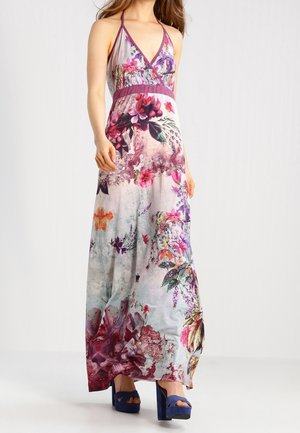 Femme portant une robe maxi fleurie sans manches avec un profond décolleté en V et des talons plateformes violets, debout devant un fond uni.