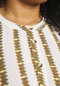 Blusa in tessuto bianco con scollatura a bottoni e motivi verticali in khaki, giallo e ruggine. Leggera con una morbida caduta.