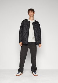 Black quilted jacket with a round collar, snap button closure, worn over a white t-shirt and paired with dark pants and white sneakers.