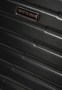 Czarna twarda walizka z teksturowaną powierzchnią z poziomymi rowkami oraz logo marki w złotym akcentem.