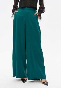 Szerokie spodnie w głębokim odcieniu teal, wykonane z g�ładkiego materiału, z wysokim stanem i kieszeniami po bokach, zestawione z czarnymi czółenkami na spiczastym obcasie.