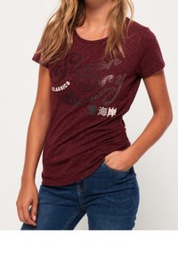 Camiseta de manga corta color burdeos con cuello redondo, que presenta un estampado gráfico texturizado en plateado y blanco, combinada con jeans azules.