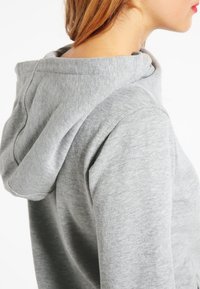 Sweat à capuche gris avec une texture douce et une capuche à cordon, présentant une coupe décontractée et des coutures décoratives aux épaules.