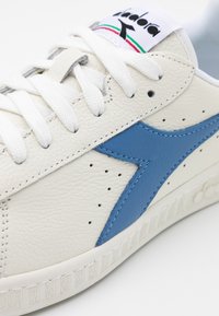 Vita lädersneakers med blå sidostrip, vita skosnören och logotyp på tungan med svart symbol och röda och gröna ränder.