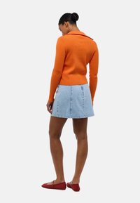 Pull orange à côtes avec un col, associé à une mini-jupe en denim bleu clair. Le modèle porte des ballerines rouges en résille et se tient les mains sur les hanches.
