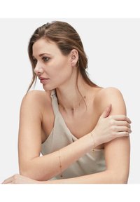 Guld smykke med delikate kædedesigns, med små vedhæng på en halskæde og armring. Båret mod en neutral stof.