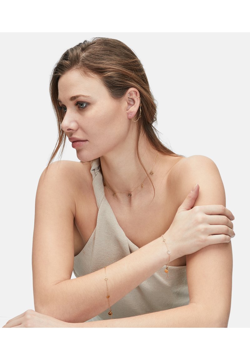 Guld smykke med delikate kædedesigns, med små vedhæng på en halskæde og armring. Båret mod en neutral stof.