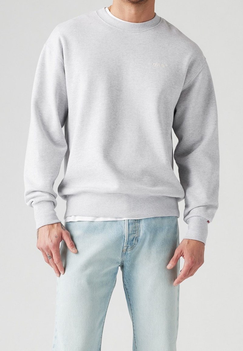 Homme portant un sweat-shirt gris clair à col rond et un jean bleu clair, debout les bras détendus le long du corps devant un fond uni.