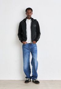 Svart zip-up jakke med logo, hvit skjorte, blå denim jeans og svarte joggesko med hvite detaljer. Modellen står mot en nøytral bakgrunn.