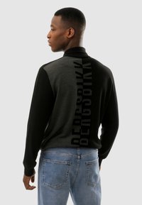 Giovane uomo che indossa un maglione nero e grigio con la scritta verticale "BERSHKA" sulla schiena, abbinato a jeans azzurri chiari, in piedi di lato.