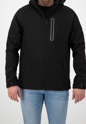Personne portant une veste noire zippée avec capuche et deux poches latérales, associée à un jean bleu clair sur un fond uni.