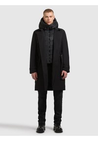 Czarny płaszcz trench z wyraźnymi ramionami, noszony na puchowej czarnej kurtce, zestawiony z czarnymi spodniami i masywnymi czarnymi botkami. Gładki materiał i elegancki design.