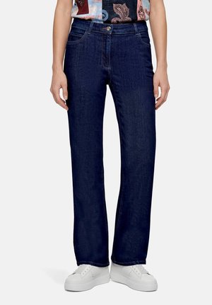 Personne portant un jean bleu foncé droit, une chemise à motifs rentrée dans le pantalon, et des baskets compensées blanches, debout sur un fond clair.