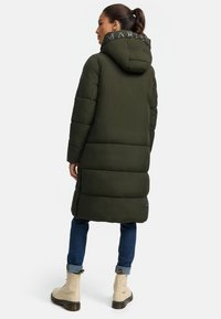 Padded olive green long coat with a hood, featuring a logo accent on the collar and a relaxed fit. Paired with light beige boots and blue jeans.