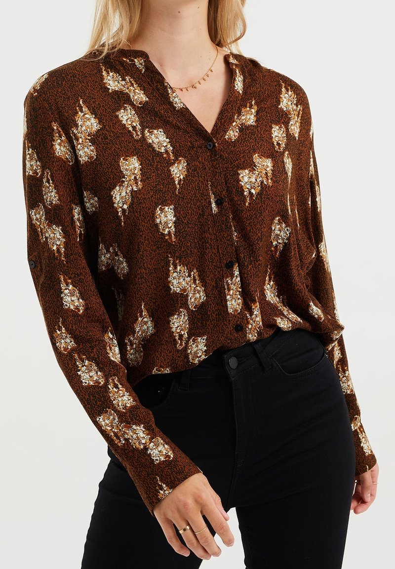 Femme portant un chemisier marron à motifs floraux boutonné, un pantalon noir et un collier en or délicat.