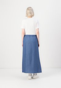 Denim-maxiskjørt med myk tekstur, elastisk midjebånd, i en mellomblå nyanse, brukt med en hvit t-skjorte og hvite sneakers.