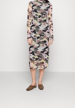 Femme portant une robe ajustée à manches longues, longueur midi, avec un motif camouflage pastel et des chaussures marron ouvertes à enfiler.