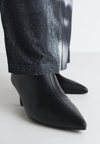 Črne usnjene gležnjarje s koničastim nosom in nizko peto, ki imajo gladko teksturo in eleganten dizajn, dopolnjene z hlačami z abstraktnimi sivimi vzorci.