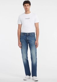 Bijela majica s logotipom "GUESS JEANS", uparena s plavim trapericama ravnog kroja, svijetlog izbljeđenja, nosi se s bijelim tenisicama.