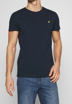 Homme portant un t-shirt bleu marine ajusté avec un petit logo oiseau jaune sur la poitrine et un jean bleu, debout devant un fond uni.