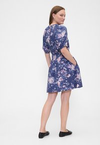 Blumenkleid in Marineblau mit kurzen Puffärmeln, elastischer Taille und seitlichen Taschen. Verziert mit rosa und violetten Blumenmustern durchgehend.