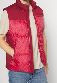 Mężczyzna w czerwonym kamizelce puchowej z geometrycznym wzorem, noszący beżowy T-shirt i niebieskie dżinsy, z jedną ręką w kieszeni kamizelki.
